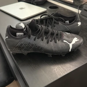 Svarta Puma Future fotbollsskor - Säljer ett par svarta Puma Future fotbollsskor med vita detaljer och logga. Skorna har snörning, låg profil och mönstrad ovandel för extra grepp. Sulan har fasta dobbar för spel på gräs. Snygg och sportig design med texten 'Future Creator Zones' på sidan.