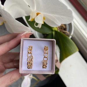 Guldiga örhängen med kedjedetalj - Snygga örhängen i guldton med kedjeliknande design och små gnistrande stenar. Perfekta för att ge din look lite extra edge och glans. Kommer i en fin ask, redo att användas direkt.