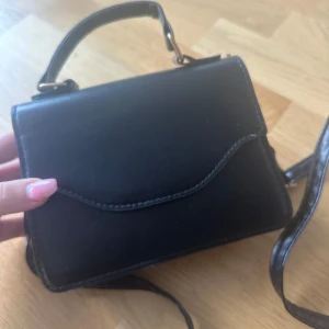 Svart axelväska i skinnimitation - En stilren svart axelväska i skinnimitation med både handtag och lång axelrem. Väskan har ett enkelt magnetlås och ett rymligt huvudfack. Perfekt för att bära det viktigaste och passar till många olika outfits.