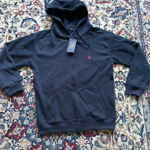 Svart hoodie från Polo Ralph Lauren - Svart hoodie från Polo Ralph Lauren med dragkedja och huva. Klassisk design med röd broderad logga på bröstet och känguruficka framtill. Tillverkad i mjuk bomull, perfekt för chill dagar. Snygg och enkel att matcha med allt.