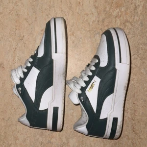 Puma sneakers i vitt och mörkgrönt - Snygga Puma sneakers i vitt och mörkgrönt med klassisk logga i guld på sidan. Skorna har snörning, rund tå och platt sula. Ovandelen är i syntet och skinnliknande material med perforerade detaljer för extra stilpoäng.