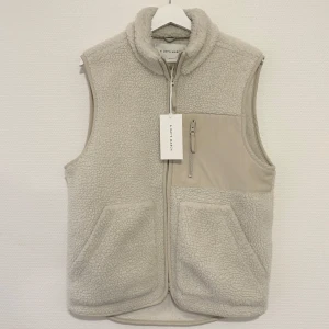 Beige fleeceväst från A Day's March - Säljer en helt ny beige fleeceväst från A Day's March med dragkedja framtill, hög krage och tre fickor, varav en bröstficka med dragkedja. Nypris 1200 kr.