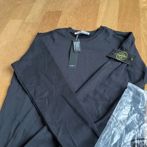 Svart sweatshirt från Stone Island - Svart sweatshirt från Stone Island med klassisk rund hals och ribbade muddar. Tröjan har den ikoniska Stone Island-loggan på vänster ärm. Perfekt för dig som gillar stilrena och trendiga plagg.