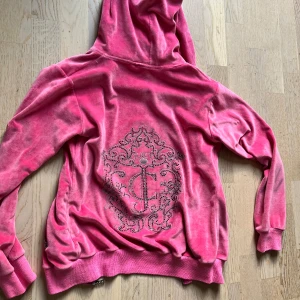 Juicy Couture rosa velourjacka - Juicy Couture hoodie i starkt rosa velour med glittrande strasstenar och ikoniskt JC-mönster på ryggen. Jackan har dragkedja och huva. Perfekt för dig som vill sticka ut med en klassisk Y2K-vibe. Köpte på loppis och kunde inte hitta lappen så vet inte exakt storlek dock skulle säga att det passar något mellan S-M