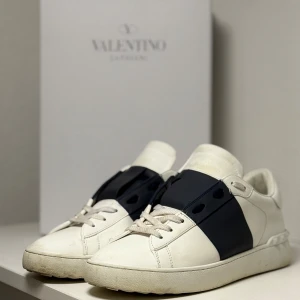 Valentino Garavani Open sneakers vit/marin - Snygga Valentino Garavani Open sneakers i vitt skinn med bred marinblå rem över mitten. Klassisk rund tå, platt sula och coola nitar baktill. Perfekta för dig som gillar exklusiva och stilrena sneakers med en twist.