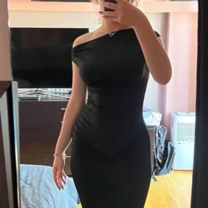 Svart festklänning - Säljer en svart långklänning med offshoulder-design och fin passform. Klänningen ger en elegant look, perfekt för dig som vill sticka ut. Materialet är stretchigt , vilket framhäver kroppen  på ett snyggt sätt.