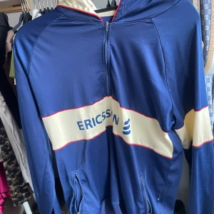 Blå Ericsson Huvtröja - Sportig blå huvtröja från Ericsson med vit och röd detalj över bröstet och ärmarna. Jackan har halv dragkedja framtill, huva och två fickor med dragkedja. Tillverkad i glansig polyester, perfekt för utomhusaktiviteter eller träning. Fläckar på baksida ärm.