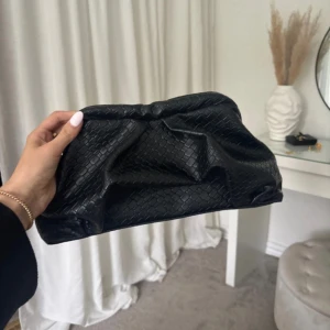 Svart kuvertväska i flätat skinn - Stilren svart kuvertväska med flätad struktur i skinnimitation. Väskan har en mjuk, puffig form och stängs med magnetlås. Invändigt finns fästen för axelrem. Perfekt att bära i handen för en trendig look.