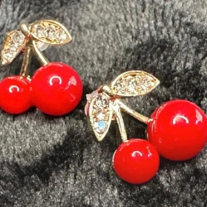 :Körsbärsörhängen i retrostil – färgglada och iögonfallande - Kawaii Rockabilly 50-tal - Lekfulla körsbärsörhängen i retrostil – perfekta för dig som gillar färg och roliga detaljer. De blanka röda körsbären är i akryl och sitter med blad i guldfärgad metall, dekorerade med små gnistrande stenar. Fästs med stift (för hål i öronen) och är helt nya. Ca 0,5 × 0,7 cm. Ingen prutning – men jag erbjuder bra rabatt vid köp av flera saker. Jag rensar smyckesskrin och garderob – spana gärna in mina andra annonser också!  