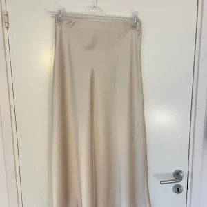 Beige satinkjol från Neo Noir - Säljer en stilren beige långkjol från Neo Noir i glansigt satinmaterial. Kjolen har en enkel och elegant design med rak passform och resår i midjan. 