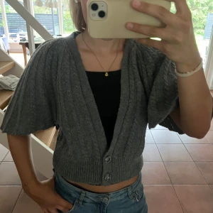 Grå sticksd bolero med vida ärmar - Så fin bolero från Massimo Dutti 💗😍Kort grå stickad kofta med bred ribbad mudd och två knappar framtill. Koftan har vida, korta ärmar och ett snyggt kabelstickat mönster. Perfekt att slänga över ett linne för en chill och trendig look. Står storlek M men skulle säga att den passar mer som S🫶🏼