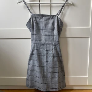 Grå rutig kortklänning Baobalei - Supersnygg grå kortklänning från Baobalei med rutigt mönster, smala axelband och öppen rygg med rosettdetalj. Klänningen har en figurnära passform och är perfekt för dig som gillar stilrena och trendiga plagg. Man knyter den själv i ryggen. 
