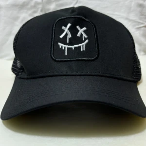 Svart truckerkeps med broderad smiley - Cool svart truckerkeps med vit broderad smiley med korsade ögon och droppande mun framtill. Kepsen har mesh-bakstycke för extra ventilation och justerbar snapback. Perfekt för dig som gillar streetstyle och vill sticka ut med en edgy detalj. Helt ny kommer i en påse och bra paketerat.