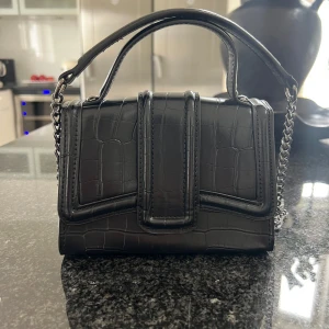 Svart axelväska med kedja och croco-mönster - Snygg svart axelväska med croco-mönstrad yta och silverfärgad kedjerem. Väskan har både handtag och lång kedja, magnetlås och stilren design. Perfekt storlek för mobil och småsaker, ger en lyxig vibe till din outfit.♥️