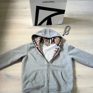 Grå Burberry hoodie med dragkedja - Säljer en grå hoodie från Burberry i bomull med klassiskt rutigt foder i huvan och insidan. Tröjan har dragkedja, broderad logga på bröstet och känguruficka. Perfekt för dig som vill ha en stilren och lyxig vardagslook.