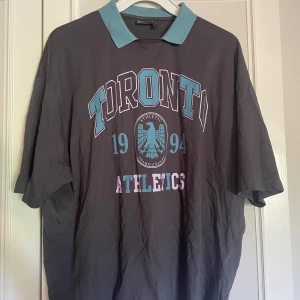 Toronto Athletics piké t-shirt - Snygg mörkgrå piké t-shirt med turkos krage och tryck i turkos och lila med texten 'Toronto Athletics 1994' och en örn på bröstet. Oversized passform och lite förbi armbåge längd på ärmar, Är i storlek M men sitter oversized så att säga så mer en L 
