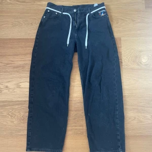 Svarta jeans från The Cava Company - Svarta jeans från The Cava Company med broderad logga på fickan och vita snören i midjan. Jeansen har raka ben och klassisk femficksdesign. Materialet är jeans och passformen är baggy.