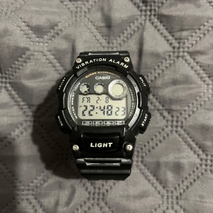 Svart digital sportklocka Casio - Digital sportklocka från Casio med svart plastarmband och rund display. Klockan har vibration alarm, super illumination, vattentålighet upp till 100 meter och flera knappar för olika funktioner. Perfekt för dig som gillar teknik och vill ha koll på tiden.