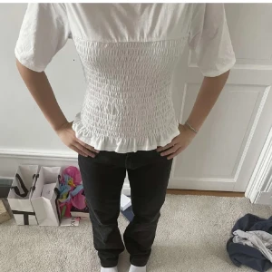 Vit smockad topp - Supersöt vit blus med smockad midja och volangkant nertill. Modellen har korta ärmar och rund halsringning. Perfekt att styla med jeans eller kjol för en clean look. Tillverkad i mjukt bomullstyg som känns skönt mot huden.