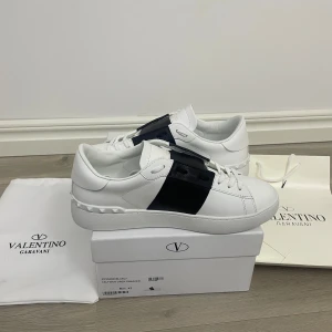 Valentino Garavani Open sneakers vit/svart - Snygga Valentino Garavani Open sneakers i vitt skinn med bred svart läderdetalj över vristen. Klassisk rund tå, vita snören och ikoniska nitar på hälen. Perfekt för dig som gillar stilrena och lyxiga sneakers med en modern twist.