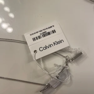 Calvin Klein halsband med pärlor och Armand  - Stilrent halsband från Calvin Klein i silverfärg med tunn kedja och minimalistisk stång som dekoreras av två vita pärlor. Perfekt för dig som gillar enkel men snygg design med en modern touch. Märkt med CK-logga på stången. Armbandet ingår oxå 