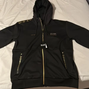Svart EA7 kofta med guld-detaljer - Svart hoodiejacka från EA7 Emporio Armani med guldiga dragkedjor och loggor. Jackan har huva med snörning, ribbad struktur och två fickor med dragkedja. Snygga guldiga detaljer på axlar och fickor ger en exklusiv vibe. Perfekt för dig som gillar streetwear med lyxig touch. Köptes igår och har aldrig använts!