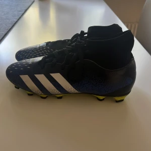 Adidas blå fotbollsskor med snörning - Säljer ett par Adidas fotbollsskor i svart och blått med vita ränder och coolt mönster på ovandelen. Skorna har snörning, högre strumpliknande skaft och gula dobbar för gräsplan. Perfekta för dig som vill sticka ut på planen!