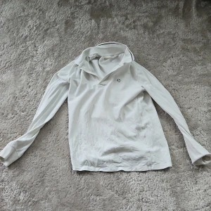 Vit långärmad pikétröja från H&M - Vit långärmad pikétröja från H&M med klassisk krage och diskret broderad detalj på bröstet. Tröjan har ribbade muddar vid ärmsluten och är gjord i mjuk bomull. Perfekt för en clean och stilren look.