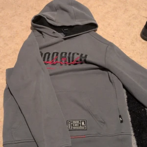Grå HOODRICH hoodie med brodyr - Snygg grå hoodie från HOODRICH med svart och röd broderad logga på bröstet. Tröjan har huva, magficka och coola detaljer som röd söm och streetwear-patch framtill. Perfekt för dig som gillar streetstyle och vill sticka ut. Den har blivit riven bara på slutet av armen 