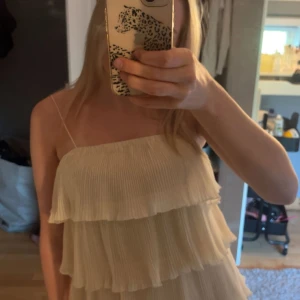 Champagnefärgad volangklänning - Supersöt kort klänning i champagnefärg med tunna axelband och flera lager volanger. Plisserad struktur ger ett luftigt och festligt intryck. Perfekt för dig som vill sticka ut med en feminin och trendig look.