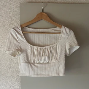 Vit ribbad croppad topp med rynk - Superfin croppad topp i vit ribbad bomull med kort ärm och fyrkantig ringning. Framsidan har snygga rynkade detaljer som ger en trendig look. Perfekt till höga jeans eller kjol för en clean och fräsch stil.❤️