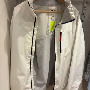 Vit vindjacka Helly Hansen - Snygg vit vindjacka från Helly Hansen i storlek L. Jackan har svart dragkedja, bröstficka med röd detalj och grå fleecefoder på insidan. Regular fit och broderad logga på insidan av kragen. Perfekt för blåsiga dagar.
