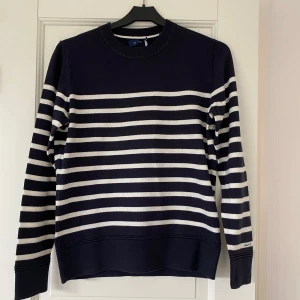 Randig marinblå tröja från GANT - Snygg marinblå tröja från GANT i storlek S med vita ränder. Klassisk rund halsringning och långa ärmar. Tröjan är i mjuk bomull och har diskret GANT-logga på ärmen. Perfekt för en stilren och avslappnad look.