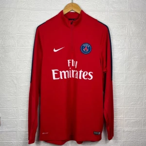 Röd PSG långärmad Nike fotbollströja - Röd långärmad fotbollströja från Nike med PSG-emblem och Fly Emirates-tryck på bröstet. Tröjan har dragkedja vid halsen och mörkblå detaljer längs axlarna. Tillverkad i 100% polyester med Dri-Fit-teknologi för bästa komfort under match eller träning.