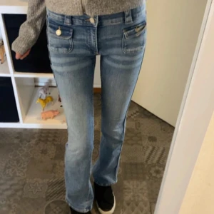 Ljusblå bootcut jeans med fickdetaljer - Säljer ett par ljusblå bootcut jeans med snygga fickdetaljer framtill och dekorativa knappar. Jeansen har låg midja och är tillverkade i klassiskt denimtyg. Perfekta för dig som gillar en avslappnad men trendig look.