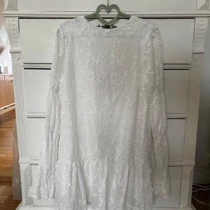 Vit blommig klänning från NA-KD - Superfin vit kortklänning från NA-KD med broderade blommor, långa ärmar och volang nertill. Klänningen har öppen rygg med knytband. Aldrig använd! 🤍