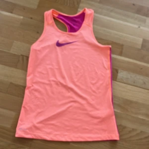 Nike Pro linne  - Nike Pro linne i neonrosa med lila detaljer och lila Swoosh-logga på bröstet. Baksidan är lila och linnet har brottarrygg. Perfekt för träning och sportiga dagar. Tillverkat i ett lätt och stretchigt syntetmaterial som andas. Storleken är barn L men är som en dam S. 