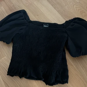 Svart smockad topp Gina Tricot S - Supersöt svart topp från Gina Tricot med smockad kropp och puffiga korta ärmar. Offshoulder-modell som ger en trendig vibe. Perfekt att styla med jeans eller kjol för en cool look.