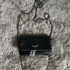 Svart axelväska med kedjerem - Snygg svart axelväska i skinn med silverfärgade detaljer och kedjerem. Väskan har dubbla dragkedjor framtill, ett coolt vingemblem i metall och extra fack på baksidan. Perfekt storlek för mobilen och det viktigaste. Stilren och edgy vibe.