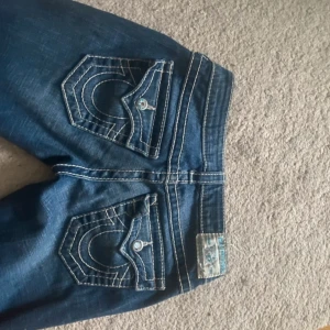 True Religion jeans med kontrastsömmar - Säljer ett par blå jeans från True Religion med tydliga vita kontrastsömmar och ikoniska fickdetaljer bak. Jeansen har låg midja och klassisk femficksdesign. Materialet är jeans med stretch för skön passform. Bootcut stil. Köpta för över 1000 i MOS, midjemått: 35cm, innerbenslängd: 80cm.
