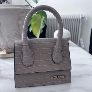 Beige Jacquemus handväska i skinn - Superfin beige handväska från Jacquemus med croco-mönstrad struktur och stilren design. Väskan har två rundade handtag och guldfärgad logga framtill. Perfekt accessoar för att lyfta din outfit.