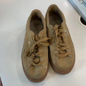 Beige Puma Suede sneakers med platåsula - Snygga beige Puma Suede sneakers med platåsula i brunt gummi. Skorna har klassisk snörning, rund tå och är tillverkade i mjuk mocka. Guldiga detaljer med Puma-logga på sidan ger en clean och stilren look. Perfekta för dig som gillar streetstyle.