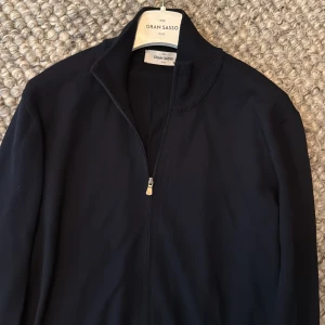 Gran Sasso tröja  - Stilren svart kofta från Gran Sasso med hel dragkedja framtill och ribbad krage. Koftan har långärmad design och är tillverkad i ett mjukt, tunt material som känns lyxigt och passar perfekt till lager-på-lager. Enkel och clean look som funkar till allt.