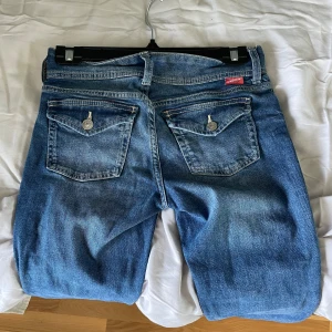 Bootcut jeans från Angel - Snygga blå bootcut jeans från Angel med klassisk femficksdesign och dubbla knappar i midjan. Jeansen har markerade bakfickor med lock och silverfärgade knappar. 