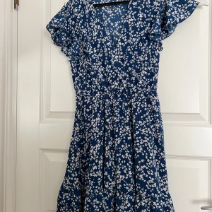 Blå blommig kortklänning med volangärm - Supersöt blå kortklänning med vita småblommor och volangärmar. Klänningen har omlott-look upptill, markerad midja och luftig kjol. Perfekt för varma dagar när du vill känna dig både chill och fin.