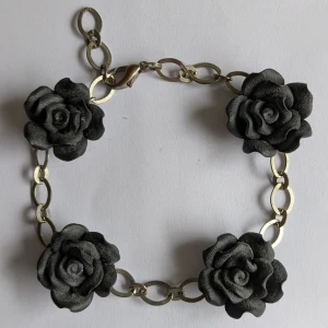 Armband med svarta rosor och kedja - Unikt armband med fyra stora svarta rosor i plast på en kedja i metallfärg. Rosorna är detaljerade och ger en cool statement-look. Kedjan har ovala länkar och ett klassiskt klolås. Perfekt för dig som vill sticka ut med accessoarer.