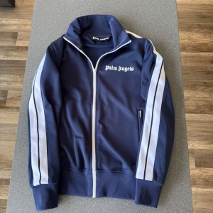 Palm Angels blå track jacket S - Säljer en blå track jacket från Palm Angels med vita ränder längs ärmarna och dragkedja framtill. Jackan har hög krage, ribbade muddar och logga på bröstet. Tillverkad i 100% polyester för en sportig vibe. Perfekt för dig som gillar streetwear och snygga detaljer. Hör av dig i dm vid frågor!