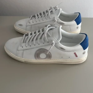 Oliver Cabell Low 1 COWBOY  - Snygga vita sneakers från Oliver Cabell med blå hälkappa och coola grå och röda detaljer på sidorna. Skorna har snörning, rund tå och är gjorda i skinn. Perfekta för dig som gillar stilrena men unika sneakers med en twist. Oliver cabellsen är sprillans nya och inte använda alls pågrund av att skorna inte passade mig 