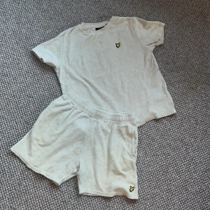 Ett lyle och scott kit med T-shirt och shorts  - Beige t-shirt från Lyle & Scott i mjuk frotté med klassisk rund hals och korta ärmar. På bröstet sitter den ikoniska gula loggan. Perfekt för chill dagar eller sommarhäng. Matcha med shorts för en avslappnad look.