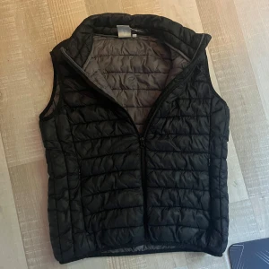 Svart dunväst från Hangten - Säljer en svart quiltad dunväst från Hangten med grå insida. Västen har hög krage och dragkedja framtill. Perfekt att slänga över en hoodie för extra värme. Klassisk och stilren design som funkar till det mesta. Stolek M men är mer som S skulle jag säga.
